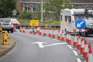 Large Quantity Of Traffoc Cones Deviding a Road, With Vehicles Driving Down One Side Of the Road. A Yellow Sign In The Background Reads "Diversion"